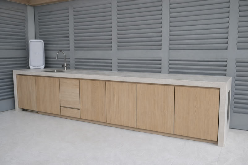 Outdoor kitchen with a long countertop, wooden cabinets, a sink, and a white appliance against a gray slatted wall.