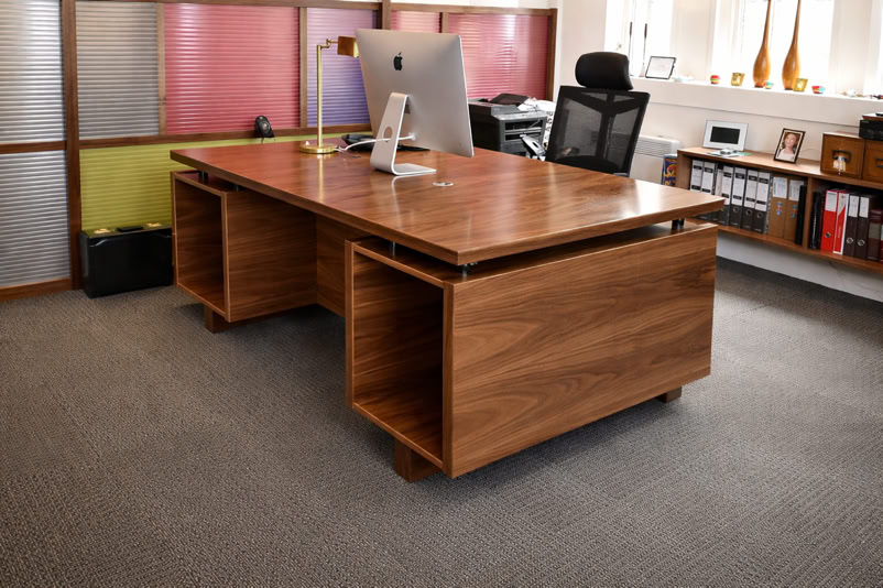 A modern office with a large wooden desk, computer, desk lamp, and office chair, surrounded by shelves and cabinets.