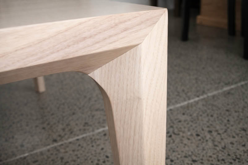 Corner of a wooden table with a smooth, angular design, set on a speckled floor.