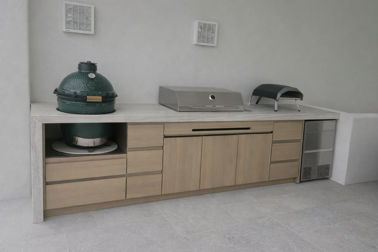 Outdoor kitchen with a green ceramic grill, a stainless steel grill, a pizza oven, and a mini fridge, set in wooden cabinetry against a light-colored wall.