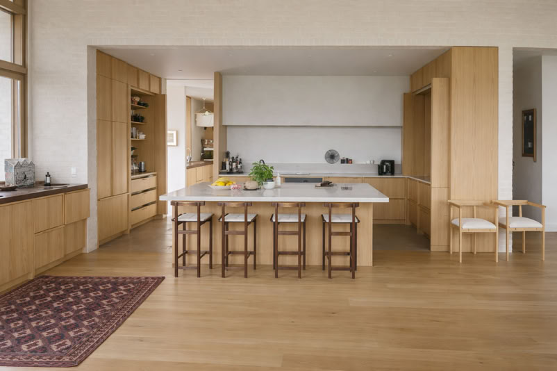 Modern kitchen with wooden cabinets, a large island with stools, and a rug on the wooden floor.