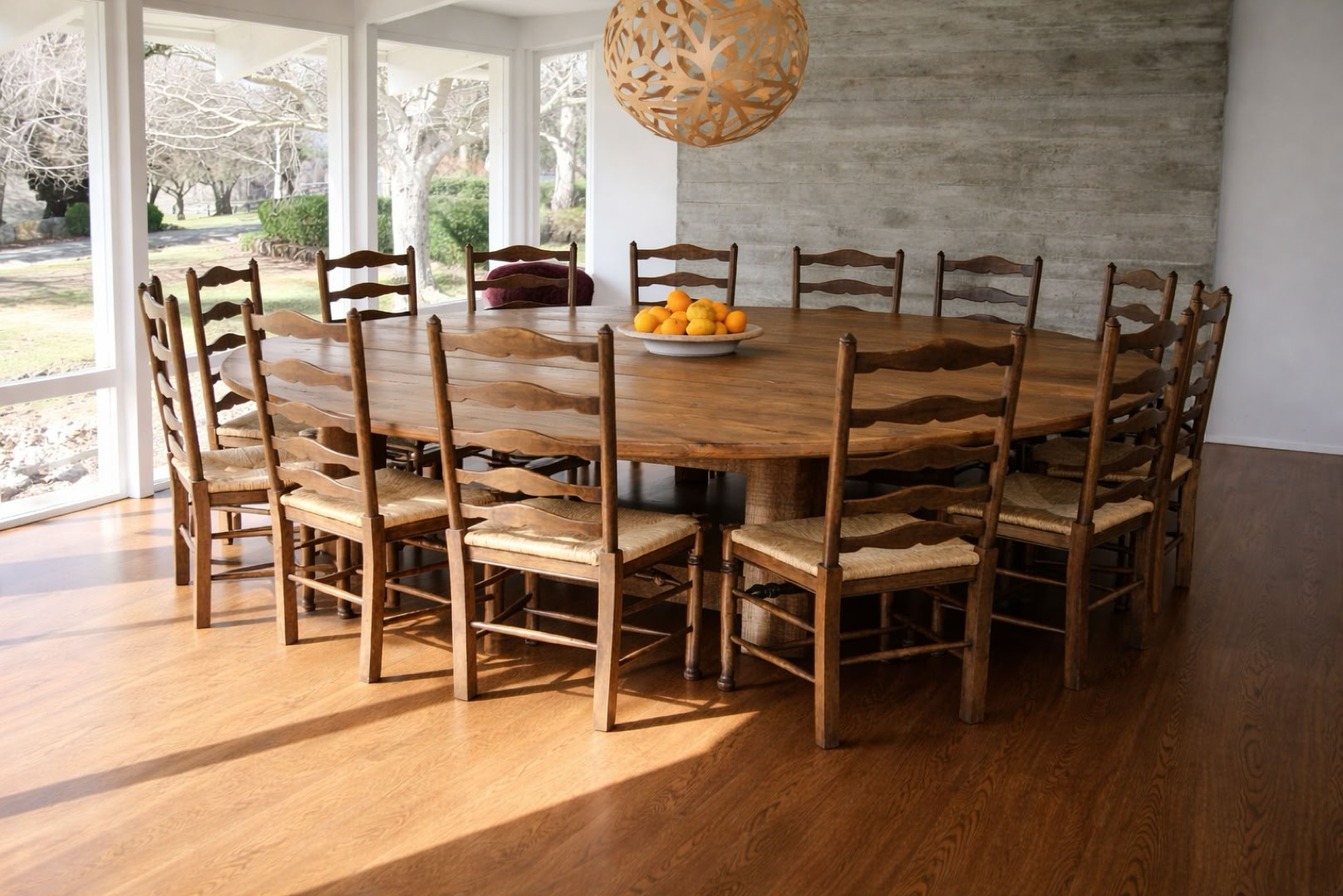 A large round wooden dining table surrounded by twelve wooden chairs with woven seats, set in a bright room with large windows and a hanging light fixture above.