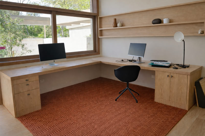A home office with a wooden L-shaped desk, two computer monitors, a black chair, a desk lamp, and shelves with decorative items. A large window and a red rug are also visible.