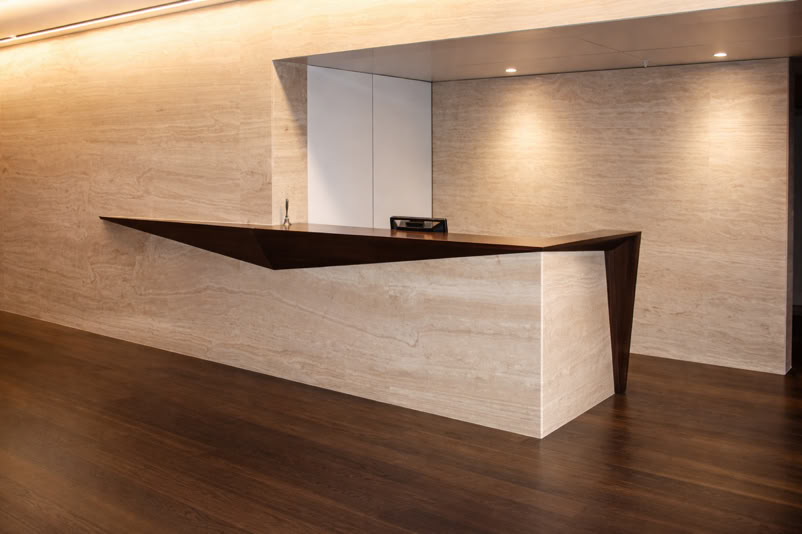 A modern reception desk with a geometric design, featuring a dark wood top and light stone base, set against a beige wall and dark wood floor.