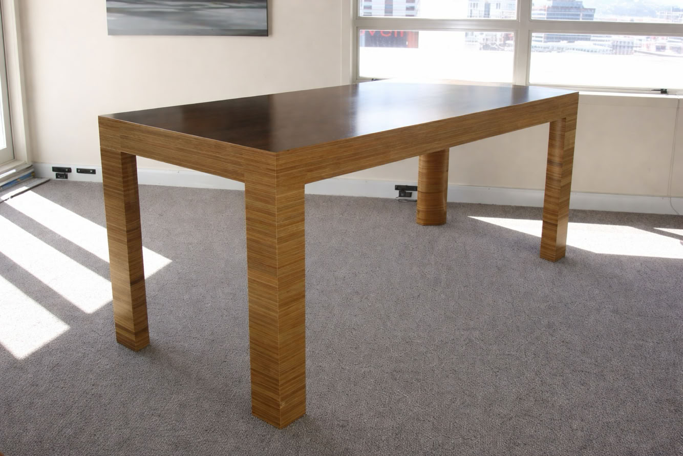 Wooden table with a dark top and light-colored legs, placed on a carpeted floor in a room with large windows.
