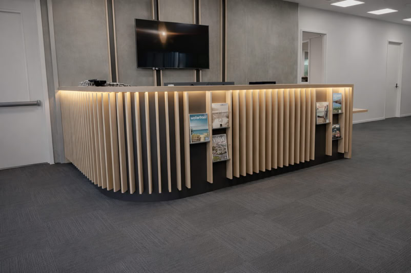 A modern reception desk with vertical wooden slats, illuminated by lights, and shelves containing magazines or brochures, set in an office environment with a wall-mounted TV above.