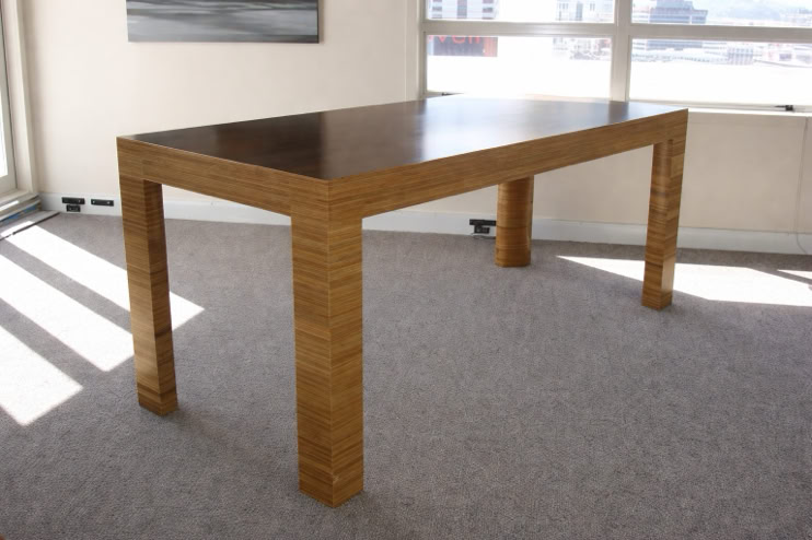 A wooden table with a dark top and light-colored legs, placed on a carpeted floor in a room with large windows.
