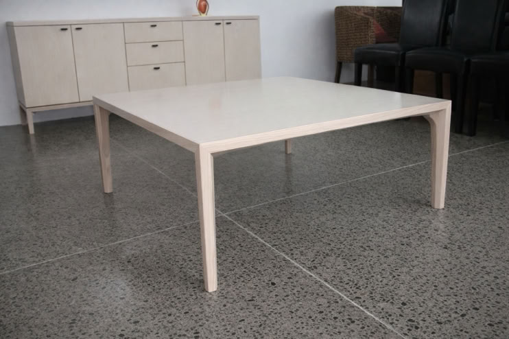 A light-colored square table on a polished concrete floor, with a beige sideboard and black chairs in the background.