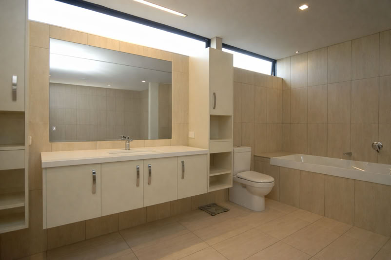 Modern bathroom with a large mirror above a double sink vanity, a toilet, and a bathtub, all with beige tile walls and floor.