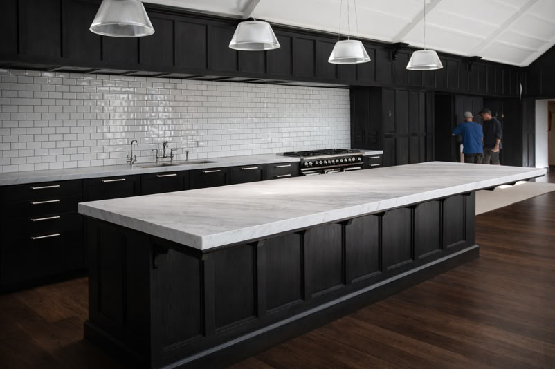 A modern kitchen with dark cabinetry, a large white marble island, stainless steel pendant lights, and a white subway tile backsplash. Two people are standing in the background.