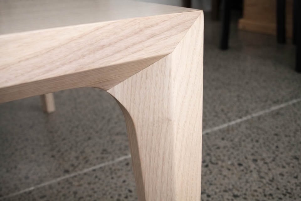 Corner of a wooden table with a smooth, angled design, set on a speckled concrete floor.