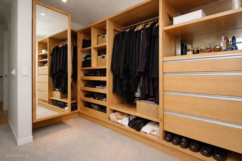 A walk-in closet with wooden shelves and drawers, containing neatly organized clothes, shoes, and accessories. A large mirror is on the left wall.