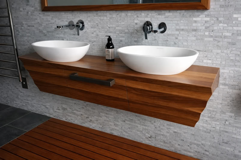 A modern bathroom with two white vessel sinks on a wooden floating vanity, a small bottle between the sinks, and a tiled wall background.
