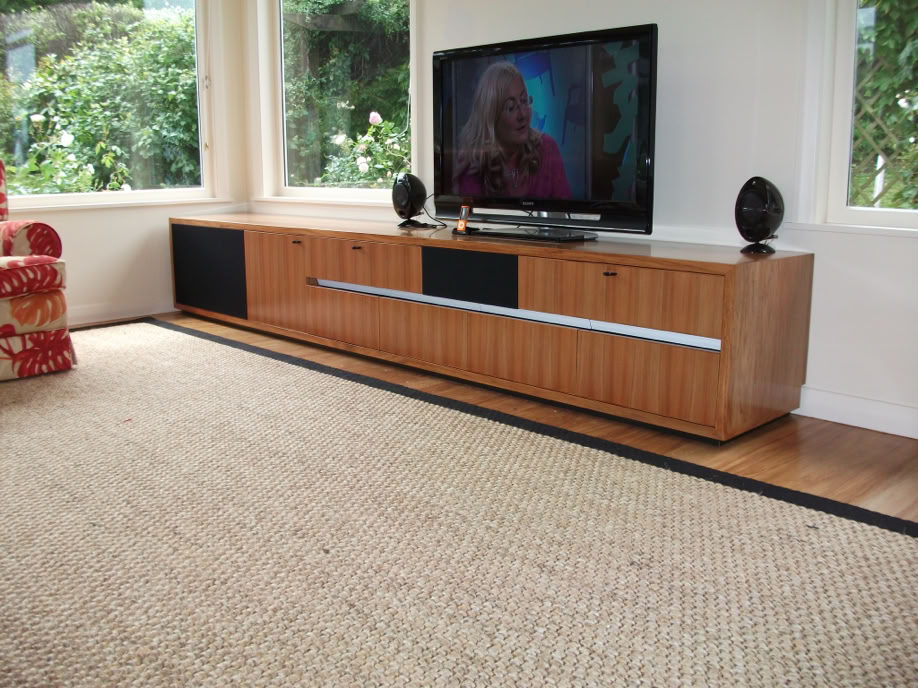 A living room with a large TV on a wooden cabinet, two black speakers, a woven rug on the floor, and a red patterned chair partially visible on the left.