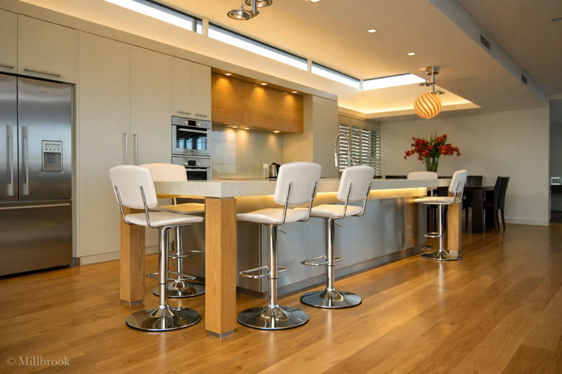 Modern kitchen with wooden flooring, a large island with four white bar stools, stainless steel appliances, and a dining area in the background.